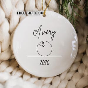 May include: White ceramic ornament with a gold string for hanging. The ornament features the name "Avery" in cursive, a bowling ball graphic, and the year "2026" in a simple font. The words "FREE GIFT BOX" are in the upper left corner.