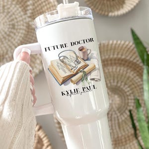 May include: White tumbler with a handle, featuring the text "FUTURE DOCTOR" and a medical-themed illustration. The illustration includes a stethoscope, open book, and a coffee cup. The name "KYLIE PAUL" is printed below the illustration.