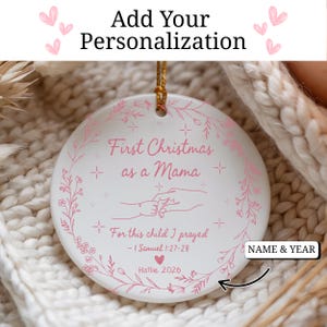 May include: A white ceramic Christmas ornament with pink text and floral accents. The ornament reads "First Christmas as a Mama" and "For this child I prayed." Includes space for personalization of name and year. A gold string is attached for hanging.