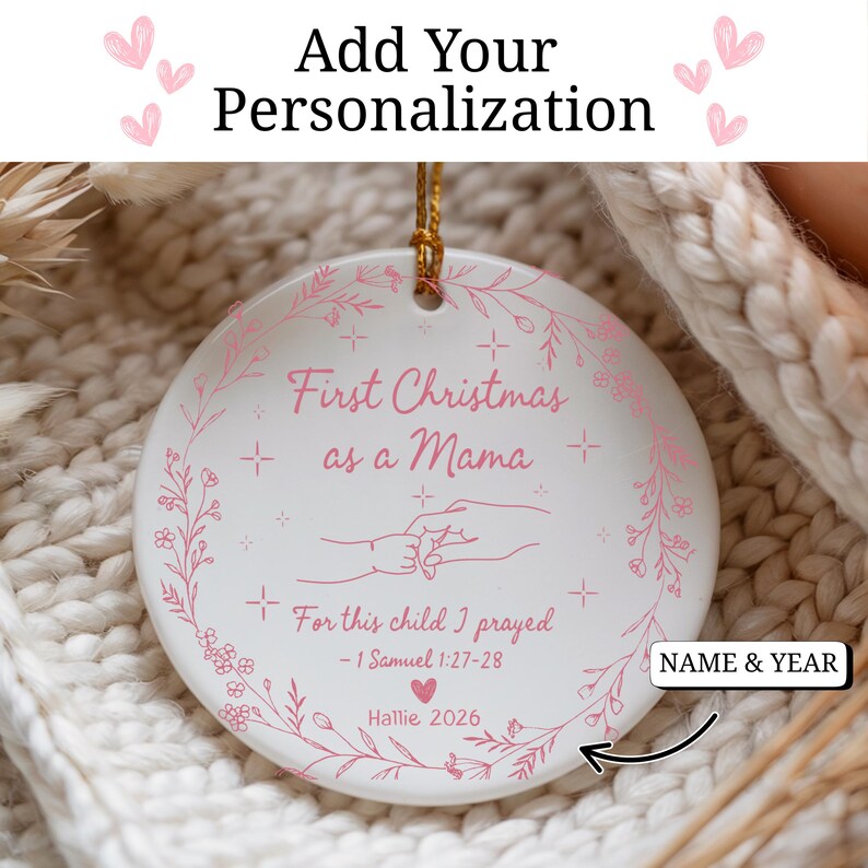 May include: A white ceramic Christmas ornament with pink text and floral accents. The ornament reads "First Christmas as a Mama" and "For this child I prayed." Includes space for personalization of name and year. A gold string is attached for hanging.