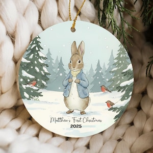 May include: A round Christmas ornament featuring a watercolor illustration of a rabbit in a blue jacket, standing in a snowy forest. The text "Matthew's First Christmas 2025" is printed at the bottom. A gold string is attached for hanging.