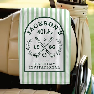 May include: A personalized golf towel with a green and white striped background. The towel features a dark green emblem with the text "Jackson's 40th Birthday Invitational" and crossed golf clubs. The years 1986 are also displayed.