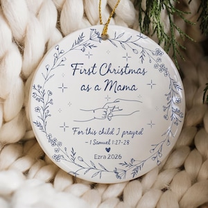 May include: A white ceramic Christmas ornament with a gold string for hanging. The ornament features the text "First Christmas as a Mama" and "For this child I prayed - 1 Samuel 1:27-28". It includes a floral design and the name "Ezra 2026".