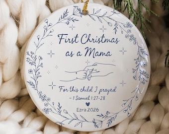 May include: A white ceramic Christmas ornament with a gold string for hanging. The ornament features the text "First Christmas as a Mama" and "For this child I prayed - 1 Samuel 1:27-28". It includes a floral design and the name "Ezra 2026".