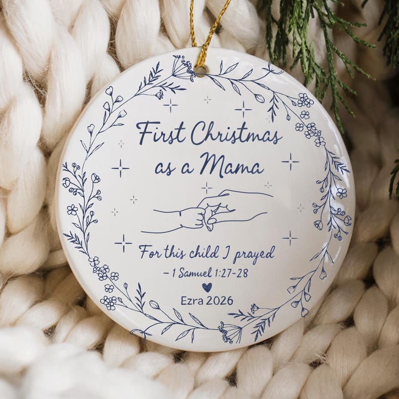 May include: A white ceramic Christmas ornament with a gold string for hanging. The ornament features the text "First Christmas as a Mama" and "For this child I prayed - 1 Samuel 1:27-28". It includes a floral design and the name "Ezra 2026".