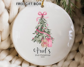 May include: White ceramic Christmas ornament with a watercolor design of a Christmas tree, pink ribbon, and a gift box. The ornament has the text "Ariel's 2nd Christmas 2025". A gold string is attached for hanging.