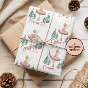 May include: Gift-wrapped package with ballerina-themed wrapping paper. The paper features illustrations of ballerinas in pink tutus, Christmas trees, and the name "Emma" in cursive. Tied with twine, the package is on a white surface with pinecones.