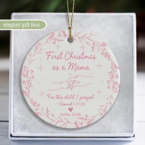 May include: A white ceramic Christmas ornament with pink text and floral accents. The ornament reads "First Christmas as a Mama" and includes the text "For this child I prayed - 1 Samuel 1:27-28". The ornament is in an elegant gift box.