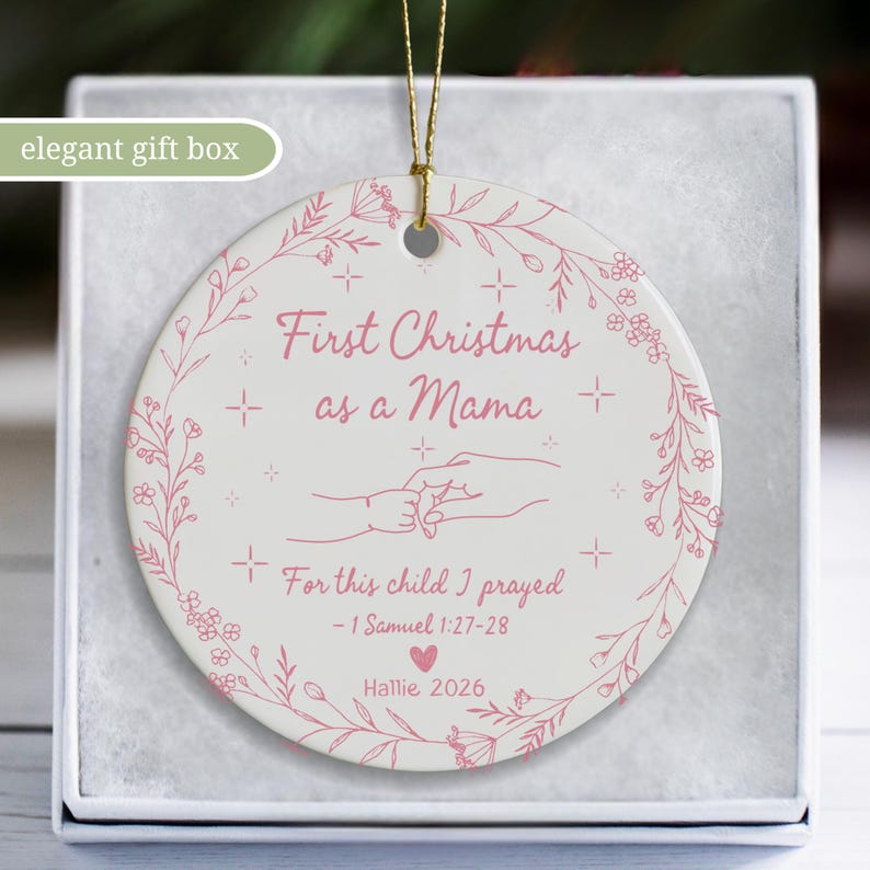 May include: A white ceramic Christmas ornament with pink text and floral accents. The ornament reads "First Christmas as a Mama" and includes the text "For this child I prayed - 1 Samuel 1:27-28". The ornament is in an elegant gift box.