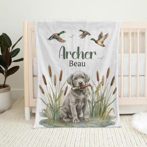 May include: A personalized baby blanket featuring a watercolor illustration of a gray Labrador puppy holding a stick, surrounded by cattails and flying ducks. The blanket has the name "Archer Beau" in green script.