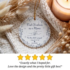 May include: A round, white Christmas ornament with the text "First Christmas as a Mama" in a floral wreath design. The ornament also includes the text "For this child I prayed - 1 Samuel 1:27-28 Ezra 2026". A gold string is attached for hanging.
