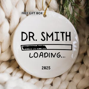 May include: A white ceramic Christmas ornament with the text "DR. SMITH LOADING... 2025" in black. The ornament has a gold string for hanging and is set against a cozy, textured background.