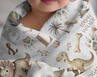 May include: A white baby blanket with a dinosaur theme. The blanket features brown dinosaurs, palm trees, bones, and footprints. The blanket is personalized with the name "Logan Walker" and the letter "L".