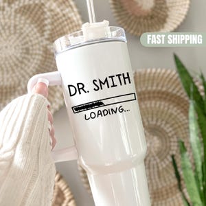 May include: White tumbler with a handle, clear lid, and a straw. The tumbler has the text "DR. SMITH" above a loading bar graphic with "LOADING..." below. The tumbler is held by a person wearing a white sweater.
