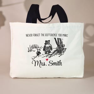 May include: A white tote bag with black handles featuring a black and white illustration of Winnie the Pooh and Piglet. The text "NEVER FORGET THE DIFFERENCE YOU MAKE" is above the illustration, with "Mrs. Smith" below.