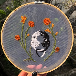 May include: Embroidered hoop art featuring two stylized cats, one black and one white, embracing. Orange flowers and green stems surround the cats. The embroidery is set against a sheer, gray fabric within a wooden hoop.
