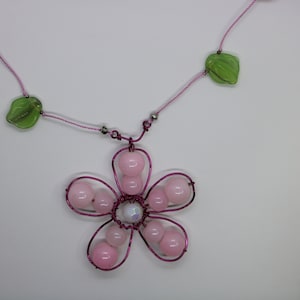 Puede incluir: Un collar de cuentas rosas y verdes con un colgante de flor rosa. La flor está hecha de alambre y tiene cuentas rosas alrededor de los bordes. El centro de la flor tiene una cuenta blanca.