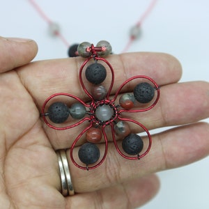 Puede incluir: Pendiente de flor envuelto en alambre rojo con cuentas de lava negra y ágata roja y gris.