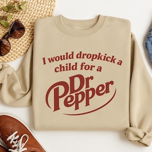 May include: Beige sweatshirt with the text "I would dropkick a child for a Dr Pepper" in maroon. The Dr Pepper logo is prominently displayed below the text. The sweatshirt is folded, with a pair of sunglasses and a brown shoe visible in the background.