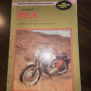 May include: A vintage book cover featuring a BSA motorcycle on a grassy field. The cover is olive green with red and pink accents. Text includes "CLYMER BSA 500 & 650cc UNIT TWINS SERVICE REPAIR PERFORMANCE" and "HIGH PERFORMANCE HOW-TO".