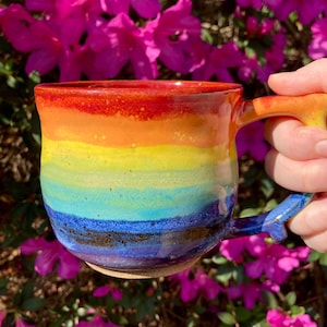 Pode incluir: Caneca de cerâmica artesanal com um design de gradiente de arco-íris. As cores transitam do vermelho na borda para o laranja, amarelo, verde, azul e roxo na base. A alça é laranja e amarela vibrantes, em um fundo de flores cor-de-rosa.