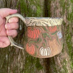 May include: A ceramic mug with a textured brown surface featuring carved and painted orange, white, and tan pumpkins with green vines. The mug has a cream-colored rim with a swirling pattern and a sturdy handle, showcasing detailed relief work.