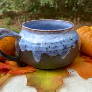 May include: A handmade ceramic mug with a dark brown glaze and a blue and white glaze on the inside. The mug has a rounded body and a handle. It is sitting on a white surface with autumn leaves.