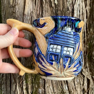 May include: A ceramic mug with a blue and tan design. The mug features a detailed depiction of a blue police box, with a tan handle and accents. The mug is held by a hand, showcasing the intricate details and craftsmanship.