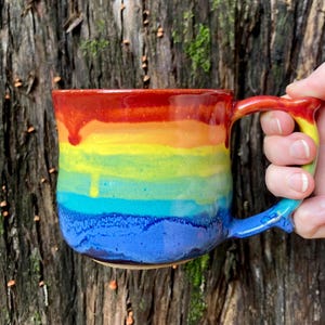 Könnte beinhalten: Eine Keramik-Tasse mit einem Regenbogen-Farbverlauf. Die Farben gehen von Rot oben nach Blau unten über, mit Gelb, Orange und Grün dazwischen. Der Griff ist ebenfalls mit den gleichen Regenbogenfarben bemalt.