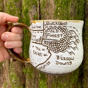 Op de afbeelding: Een witte keramische mok met een bruin, takvormig handvat. Het oppervlak is voorzien van een handgetekende kaart in donkerbruine inkt, met labels voor Buckland, Old Forest, Michel Delving, The Shire, Brandywine en Barrow Downs.