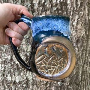 Könnte beinhalten: Eine handgefertigte Keramiktasse mit blauer und weißer Glasur. Die Tasse zeigt einen braunen Halbmond mit einem Bergketten-Design und Sternen.