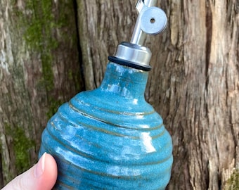 Puede incluir: Una botella de aceite de cerámica turquesa hecha a mano con una boquilla de acero inoxidable. La botella tiene un diseño en espiral texturizado y un borde oscuro. La botella de cerámica se sostiene en una mano.