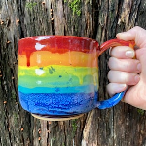 Pode incluir: Uma caneca de cerâmica artesanal com um esmalte arco-íris. A caneca apresenta um gradiente vibrante de cores, incluindo vermelho, laranja, amarelo, verde, azul e índigo. A alça também é pintada com as mesmas cores do arco-íris. A caneca é segurada por uma pessoa.