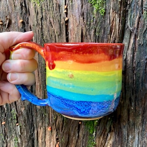 Könnte beinhalten: Eine Keramik-Tasse mit Regenbogen-Farbverlauf. Die Tasse wechselt von Rot oben nach Blau unten, mit Gelb und Grün dazwischen. Der Griff ist blau und die Tasse wird vor einem strukturierten Baumstamm gehalten.