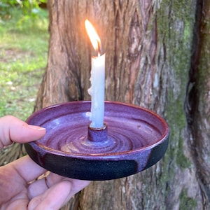May include: A round ceramic candle holder with a glossy purple interior and a matte black exterior. The holder features a central raised socket containing a thin white taper candle with a lit flame and visible wax drips running down the side.