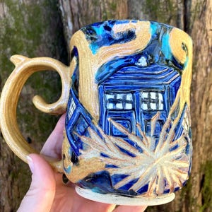 May include: A handcrafted ceramic mug with a distinctive design. The mug showcases a blue police box motif set against a tan and blue backdrop. The handle is tan and has a curved shape. A unique piece of pottery.
