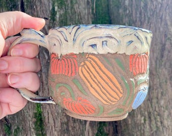 May include: Handmade ceramic mug with a brown base and a cream-colored rim. The mug features a colorful design of pumpkins, gourds, and leaves in orange, green, and blue. The handle is curved and matches the rim's color.