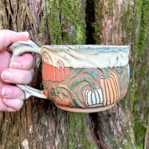 May include: A ceramic mug with a wide handle featuring a textured, carved surface depicting orange, white, and tan pumpkins with green vines. The upper rim of the mug is glazed in a smooth, creamy off-white color, while the lower body has a natural, earthy brown clay finish.