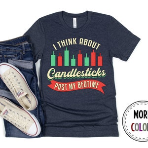 以下が含まれることがあります： ヴィンテージスタイルのフォントで「I think about candlesticks past my bedtime」と書かれたネイビーブルーのTシャツ。テキストは赤いリボンとキャンドルスティックチャートのグラフィックで囲まれています。