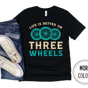 May include: Black t-shirt with the text "Life is better on three wheels" in white and a graphic of three turquoise bicycle wheels.