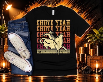 May include: A black t-shirt featuring a graphic of a cowboy riding a bucking bull with the repeated text CHUTE YEAH in yellow, orange, and red. The shirt is paired with blue denim jeans and a pair of off-white canvas sneakers.