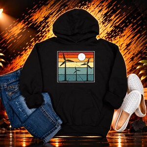 May include: A black hooded sweatshirt featuring a rectangular graphic print of three wind turbines against a sunset background with horizontal stripes in red, orange, yellow, and teal, paired with blue denim jeans and white slip-on shoes.