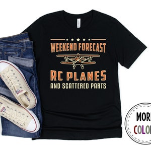 May include: Black t-shirt with the text "WEEKEND FORECAST RC PLANES AND SCATTERED PARTS" in orange and white. The shirt features a biplane graphic. A pair of beige canvas sneakers and blue jeans are next to the shirt.