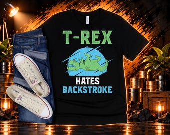 May include: A black short-sleeve t-shirt featuring a graphic of a green T-Rex dinosaur swimming on its back inside a blue circle. The text reads T-REX HATES BACKSTROKE in green and blue lettering. The shirt is paired with blue denim jeans.