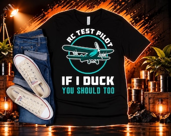 May include: A black short-sleeve t-shirt featuring a graphic of a teal airplane inside a circle with the text RC TEST PILOT and IF I DUCK YOU SHOULD TOO in white and teal lettering. The shirt is paired with blue denim jeans and white sneakers.