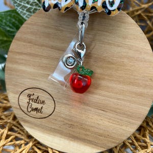 May include: A red apple charm with a green stem and leaves hangs from a silver retractable badge holder. The badge holder is attached to a leopard print charm with the word "DIVA" written in white.