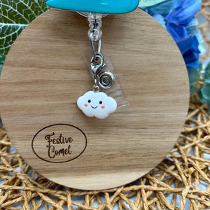 May include: A light blue badge reel with a smiling cloud charm. The reel features a stack of three blue shapes with eyes. The wooden base has the text "Festive Comet" in an oval.