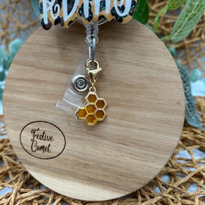 Puede incluir: Un portagafete retráctil con un diseño de estampado de leopardo, adornado con un dije de panal de abeja dorado. El portagafete está etiquetado como "Festive Comet".