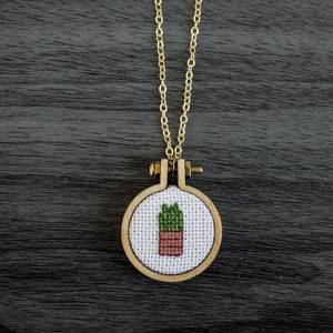 Puede incluir: Un collar de cadena dorada con un pequeño colgante circular de madera con un diseño de punto de cruz de una planta verde en maceta.