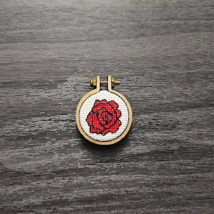 以下が含まれることがあります： 白い背景に赤いバラがクロスステッチで刺繍された、小さな丸い刺繍枠です。